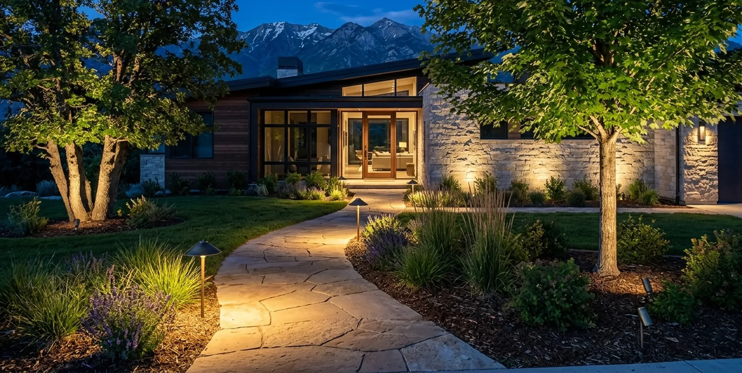Upscale mountain-modern home at dusk with uplit trees, path lights along a flagstone walk and light grazing the stone front