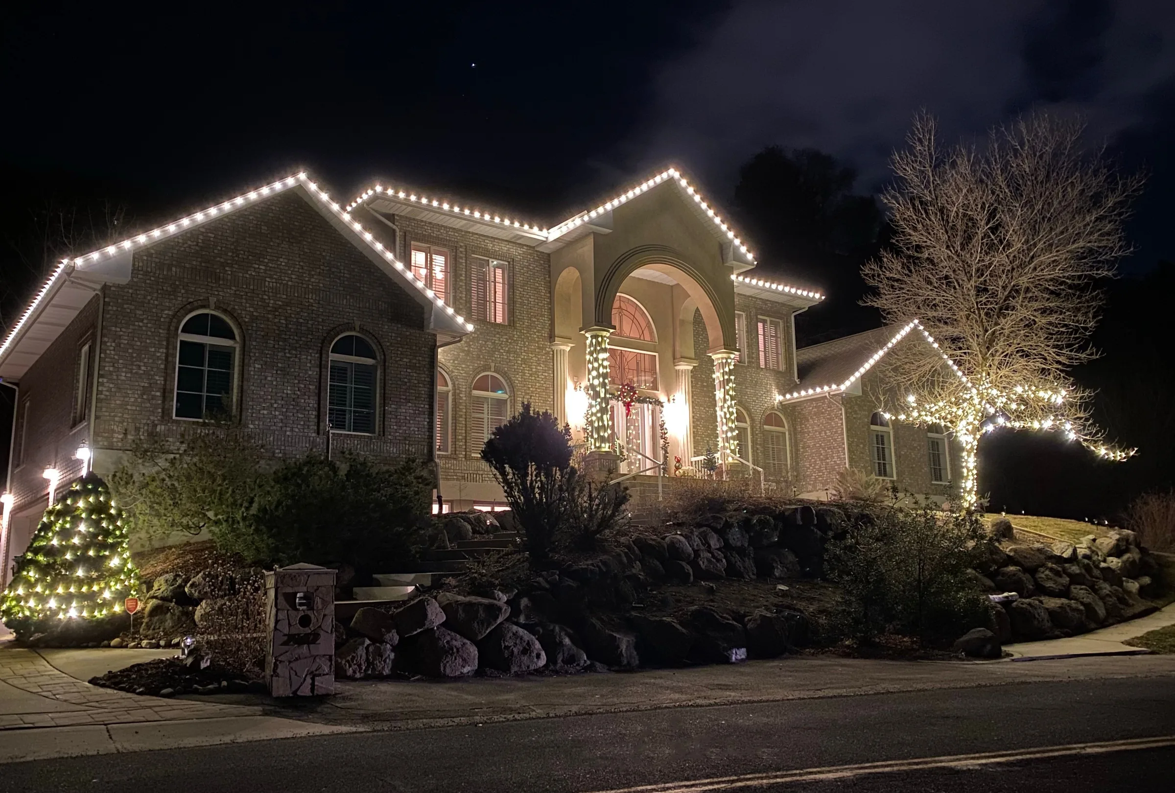 Brick home with lit rooflines and wrapped trees