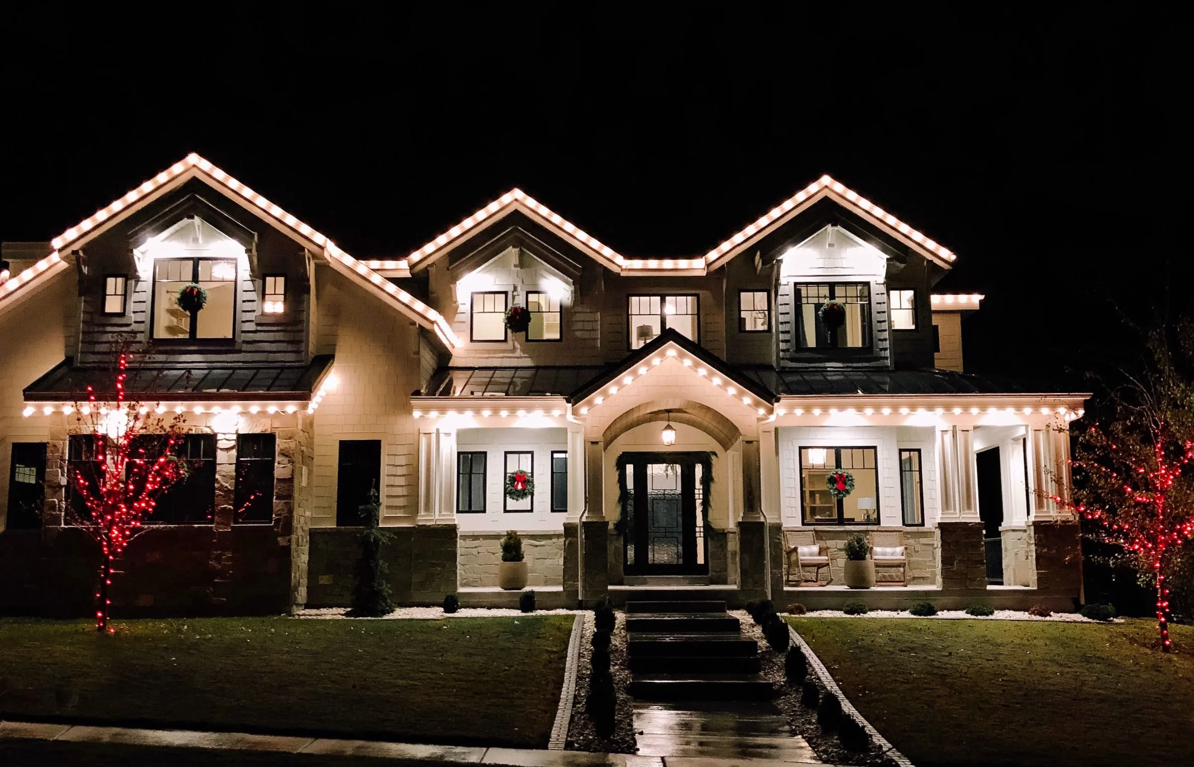 Dark craftsman home with lit gables and red wrapped trees