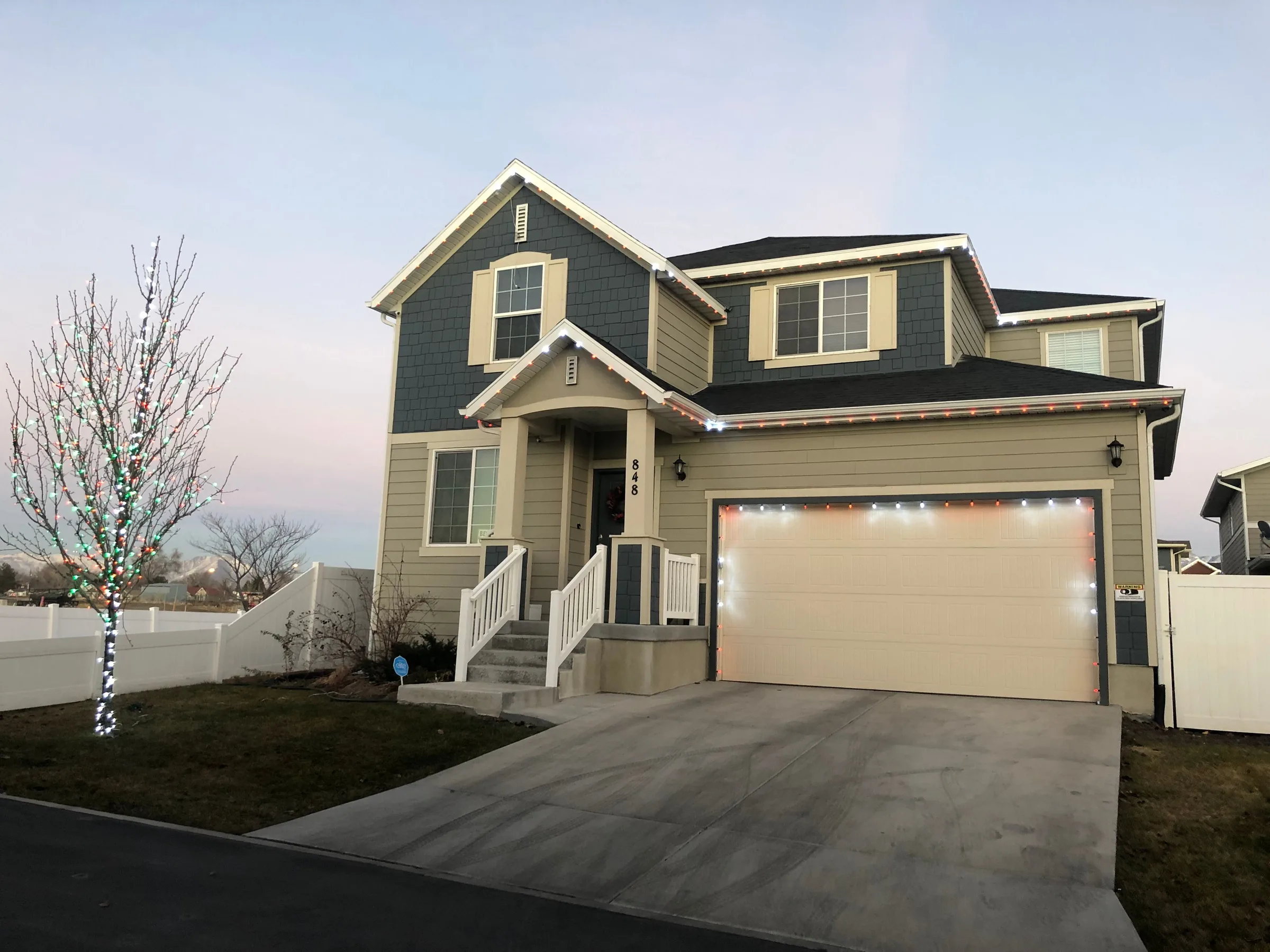 Two-story home with roofline lights and a wrapped tree at dusk